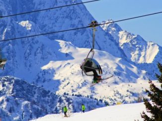 Skiers ride a chair lift above a snowy slope in the Passo del Tonale area, with the Dolomites in the background.