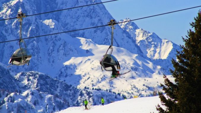 Skiers ride a chair lift above a snowy slope in the Passo del Tonale area, with the Dolomites in the background.