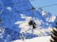 Skiers ride a chair lift above a snowy slope in the Passo del Tonale area, with the Dolomites in the background.