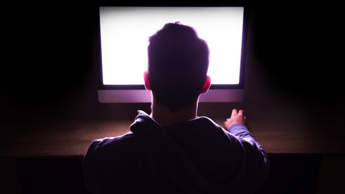 Back view of a man in front of bright computer working in the dark.