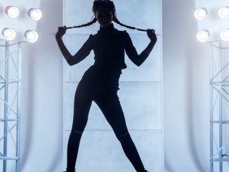 Beautiful slim young woman with two braids dressed in a black clinging clothes. The young woman dancer is performing the dance in the vogue style. She is standing holding the braids in hand. Studio shooting on a white background with light blue backlighting on a two stands