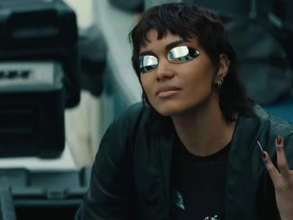 girl with short cropped dark hair wearing futuristic glasses and holding up a hand with long metallic fingernails