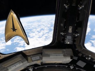 A golden triangular-shaped badge floats in microgravity aboard a space station. A blue and white planet can be seen outside the window behind the badge.