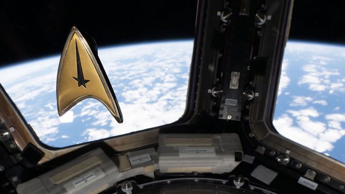 A golden triangular-shaped badge floats in microgravity aboard a space station. A blue and white planet can be seen outside the window behind the badge.