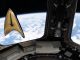 A golden triangular-shaped badge floats in microgravity aboard a space station. A blue and white planet can be seen outside the window behind the badge.